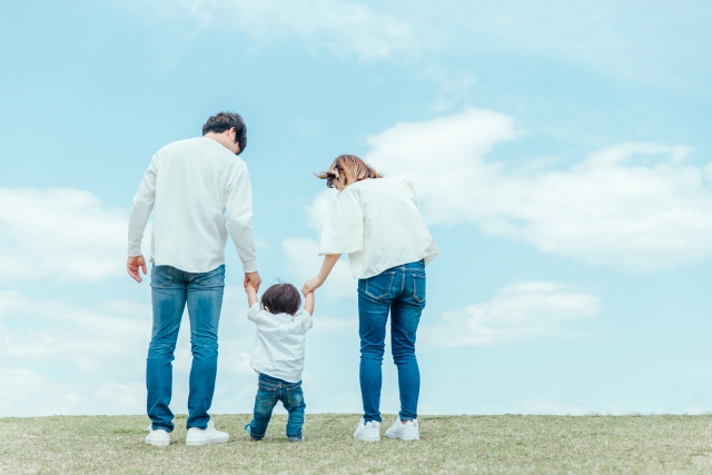 青空と芝生の上で小さな子どもと手を繋いで歩く親子の後ろ姿

